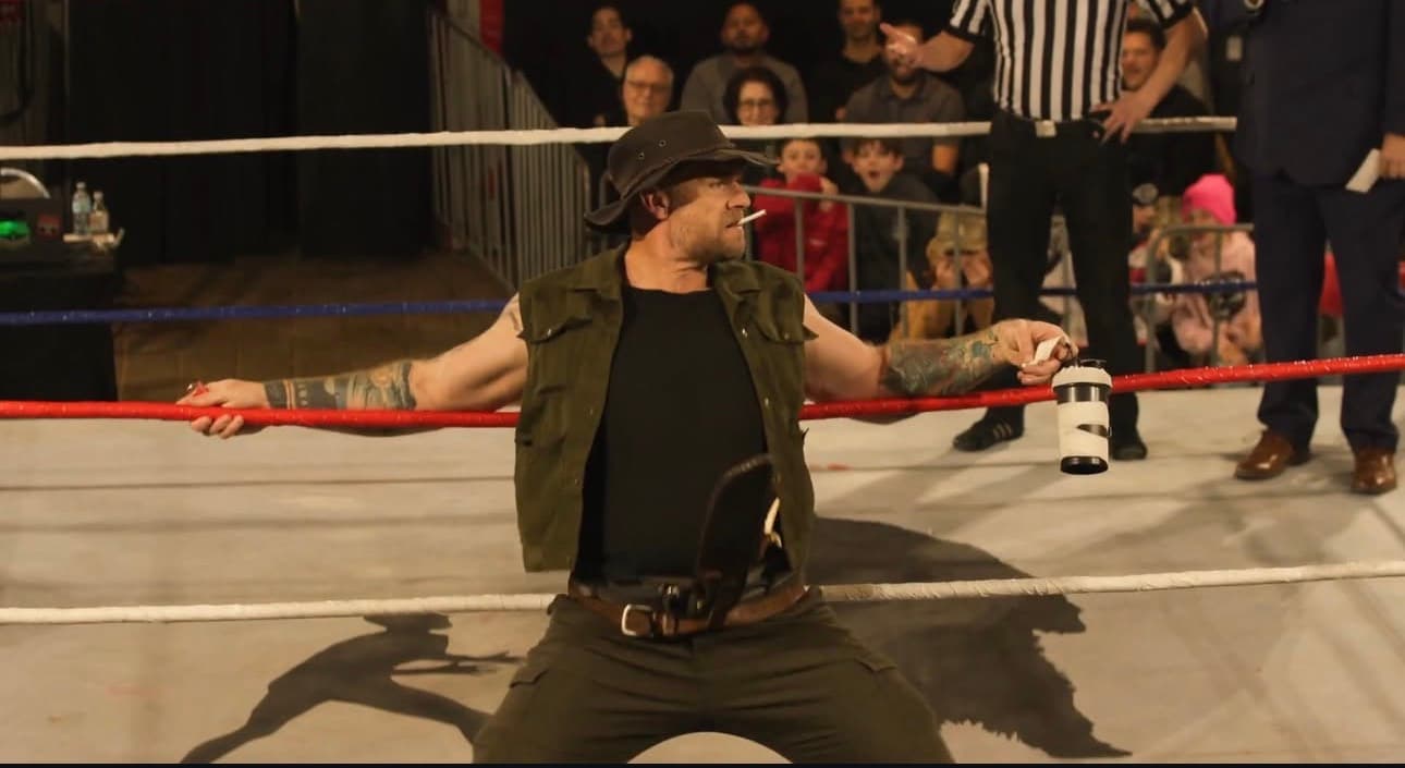 Wrestler in a fedora and green vest leans against ring ropes with a cigarette.